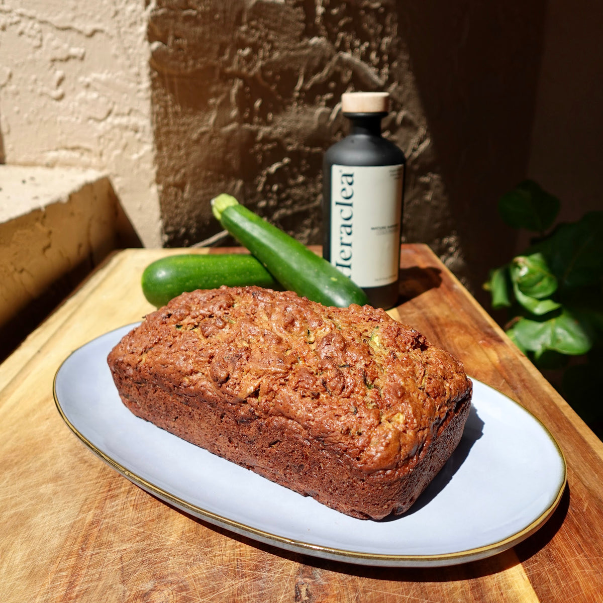 Loaf of olive oil zucchini bread on a white platter with fresh zucchini and a bottle of Heraclea Mature Harvest olive oil