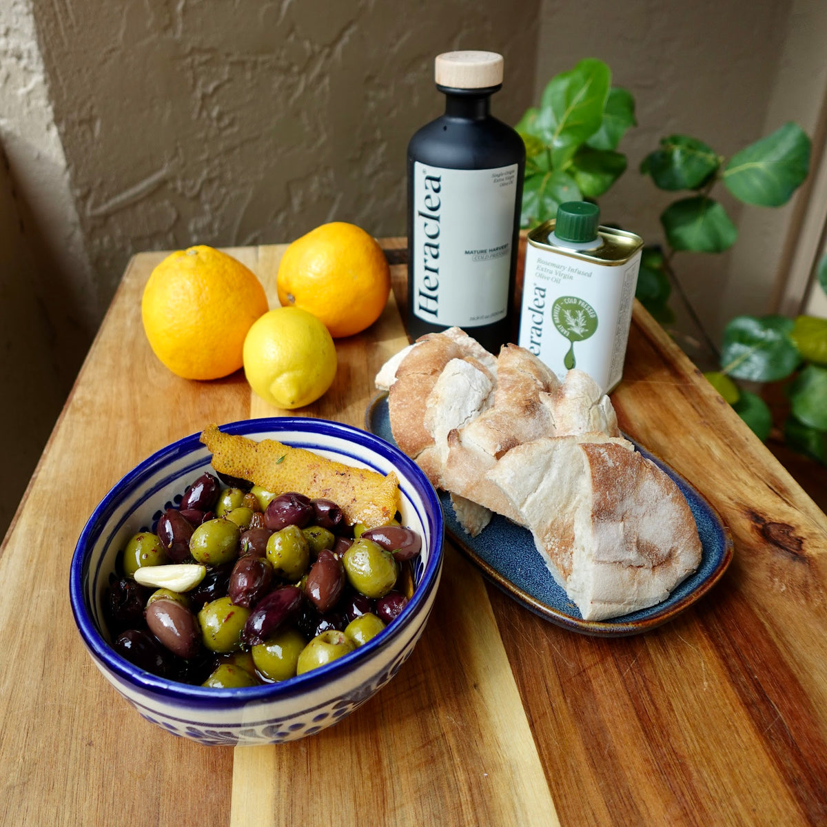 Bowl of warm marinated Kalamata and Castelvetrano olives with garlic and citrus peel, next to crusty bread, lemons and Heraclea olive oil bottles