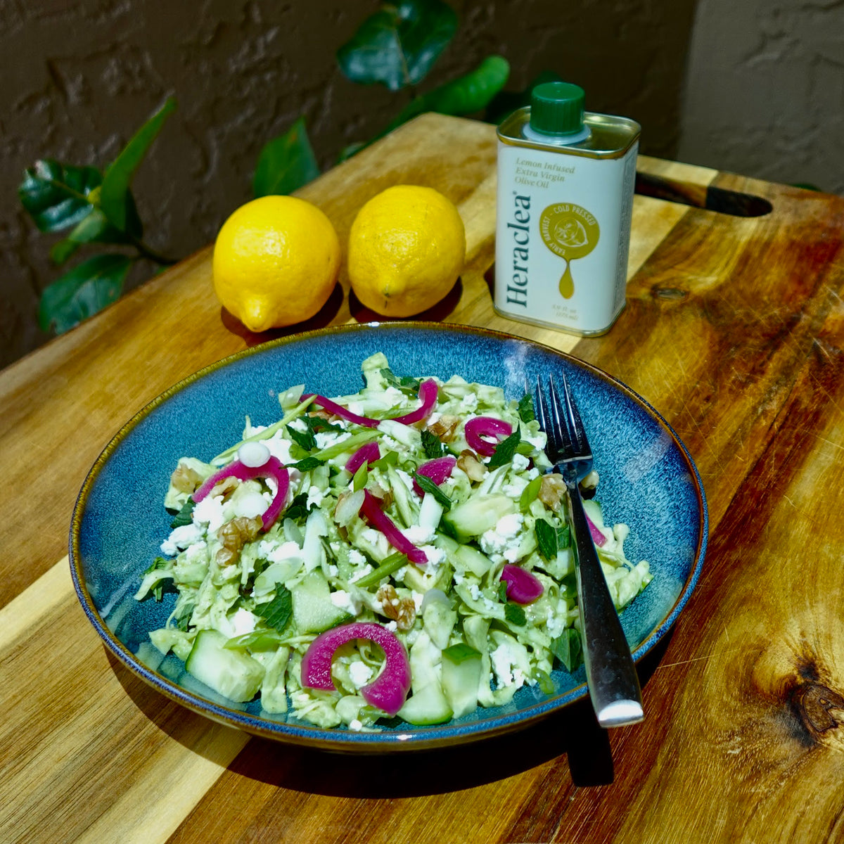 Green goddess salad of shredded cabbage, feta, cucumber, walnuts and pickled red onions on a blue plate with lemons and a tin of Heraclea Lemon Infused olive oil