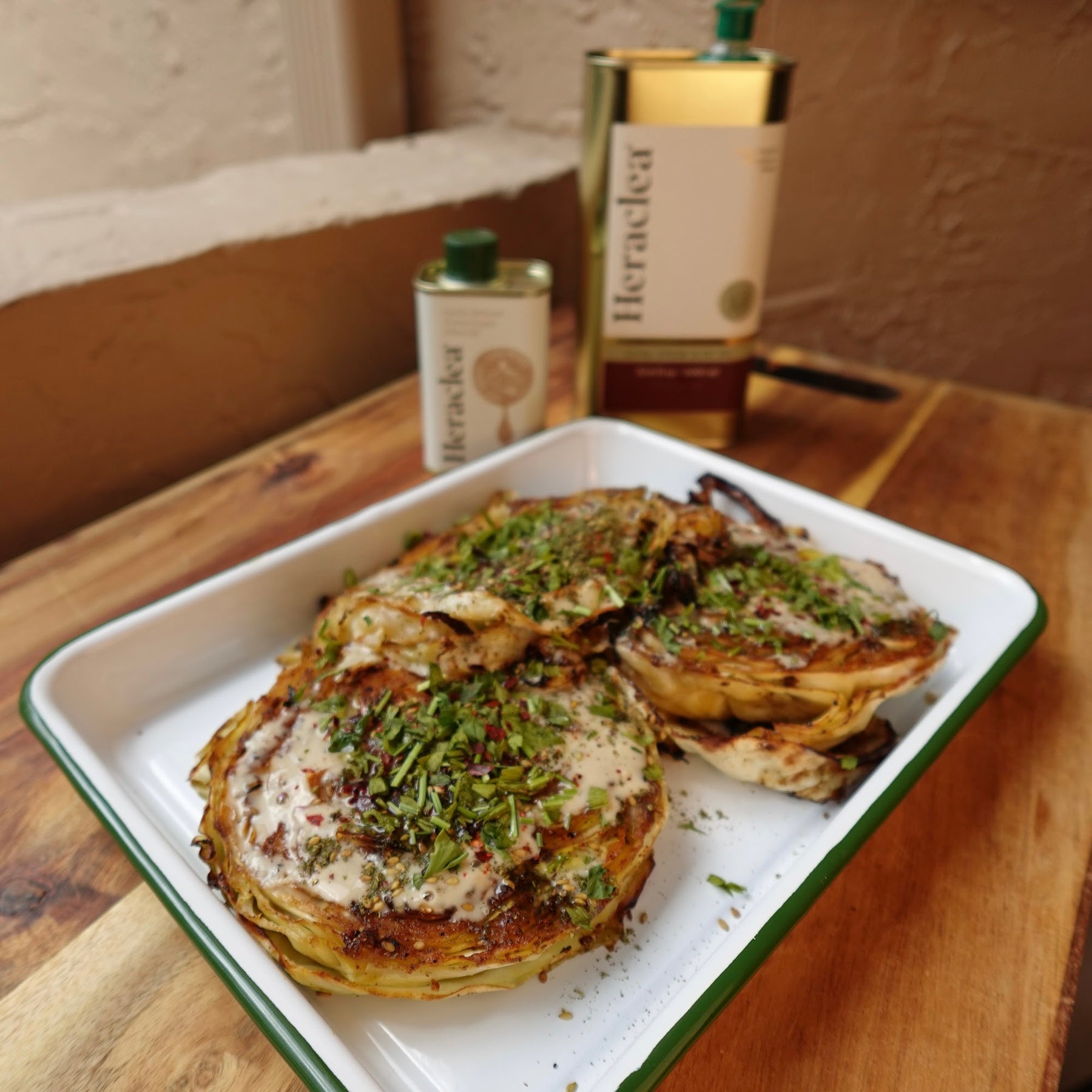 Roasted cabbage steaks with tahini-maple-garlic sauce, herbs, and za'atar on a white enamel tray next to Heraclea olive oil bottles