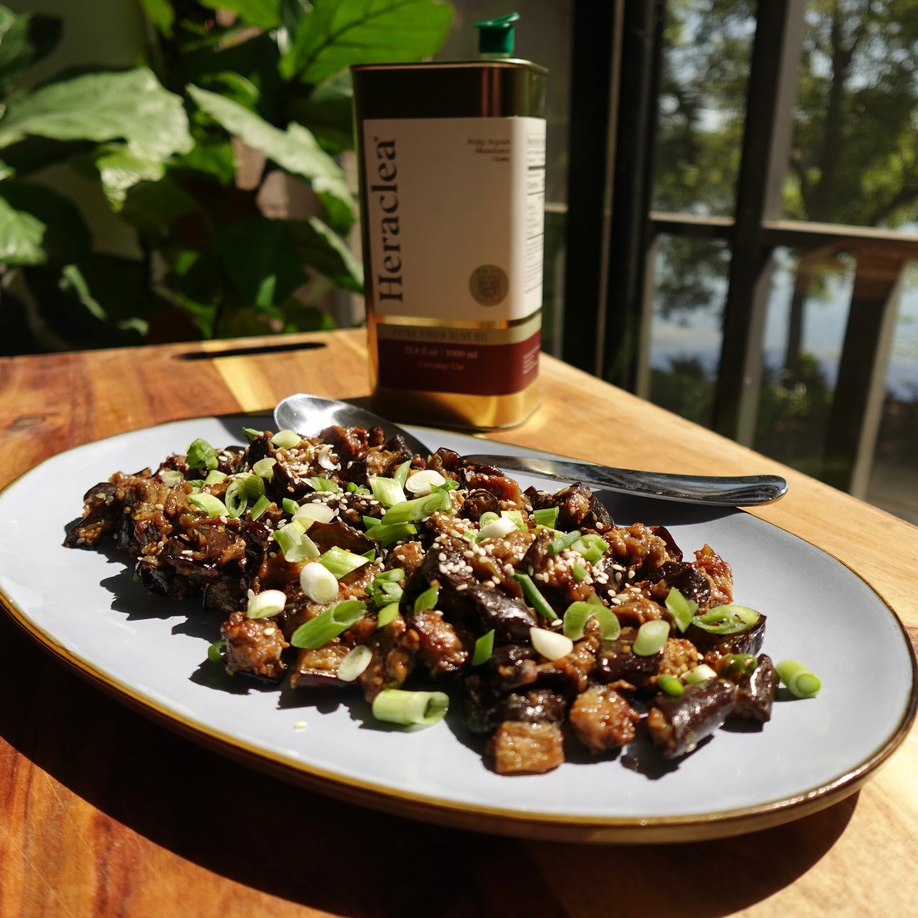 Miso glazed eggplant with sesame seeds and green onions on a gray plate next to a Heraclea olive oil bottle