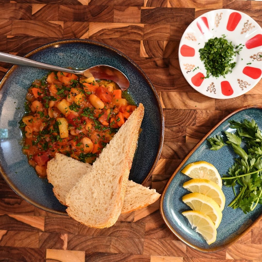 Turkish braised cannellini beans with vegetables in a blue bowl, served with bread, lemon wedges and fresh herbs