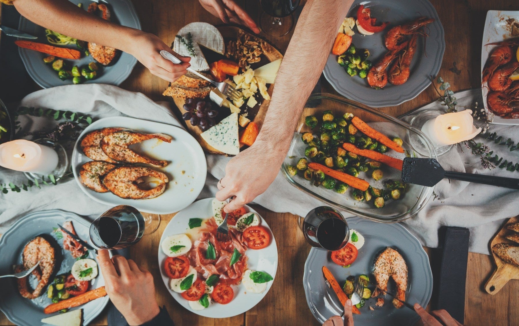 Mediterranean dinner table overhead view — salmon, brussels sprouts with carrots, caprese with prosciutto, cheese, shrimp, red wine — the kind of spread where olive oil is the default cooking fat